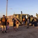 A group of civilians, including children, are gathered around a military tactical vehicle (appears to be a Joint Light Tactical Vehicle or similar JLTV/MRAP-type vehicle) in an arid, desert environment. One individual in military camouflage uniform appears to be conducting a demonstration or tour of the vehicle. The setting suggests a public military open house or community outreach event, likely at or near a military installation.