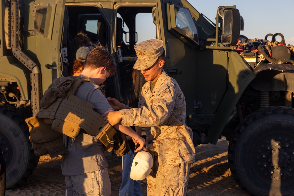 A U.S. Marine in desert MARPAT camouflage uniform is helping a young civilian child try on a tactical plate carrier vest in front of a military tactical vehicle. The scene appears to take place during a public outreach event, air show, or base open house, given the informal and educational nature of the interaction. Warm golden-hour lighting suggests the event is taking place in the late afternoon.