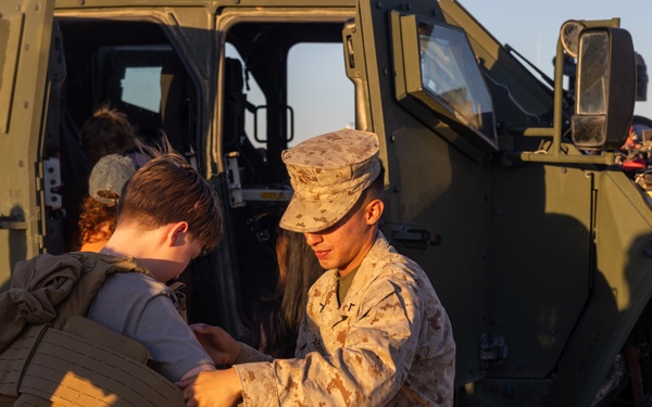 A U.S. Marine in desert MARPAT camouflage uniform is helping a young civilian child try on a tactical plate carrier vest in front of a military tactical vehicle. The scene appears to take place during a public outreach event, air show, or base open house, given the informal and educational nature of the interaction. Warm golden-hour lighting suggests the event is taking place in the late afternoon.