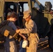 A U.S. Marine in desert MARPAT camouflage uniform is helping a young civilian child try on a tactical plate carrier vest in front of a military tactical vehicle. The scene appears to take place during a public outreach event, air show, or base open house, given the informal and educational nature of the interaction. Warm golden-hour lighting suggests the event is taking place in the late afternoon.