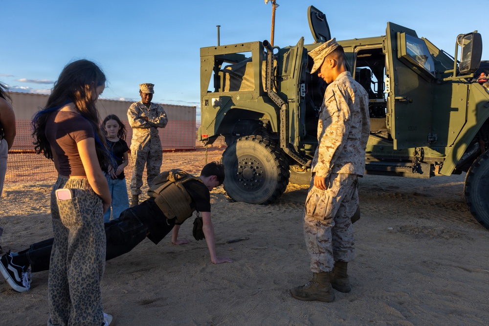 A military demonstration or public outreach event where a civilian is performing push-ups on sandy ground while wearing a tactical vest, supervised by a Marine in desert camouflage uniform. Other civilians, including a young girl and a woman in leopard-print pants, observe alongside another Marine standing in the background. A large olive-drab military tactical vehicle (likely a JLTV or similar) is parked in the background.