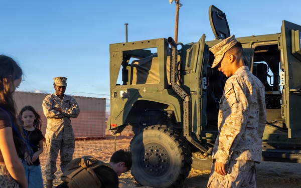 A military demonstration or public outreach event where a civilian is performing push-ups on sandy ground while wearing a tactical vest, supervised by a Marine in desert camouflage uniform. Other civilians, including a young girl and a woman in leopard-print pants, observe alongside another Marine standing in the background. A large olive-drab military tactical vehicle (likely a JLTV or similar) is parked in the background.