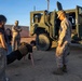 A military demonstration or public outreach event where a civilian is performing push-ups on sandy ground while wearing a tactical vest, supervised by a Marine in desert camouflage uniform. Other civilians, including a young girl and a woman in leopard-print pants, observe alongside another Marine standing in the background. A large olive-drab military tactical vehicle (likely a JLTV or similar) is parked in the background.