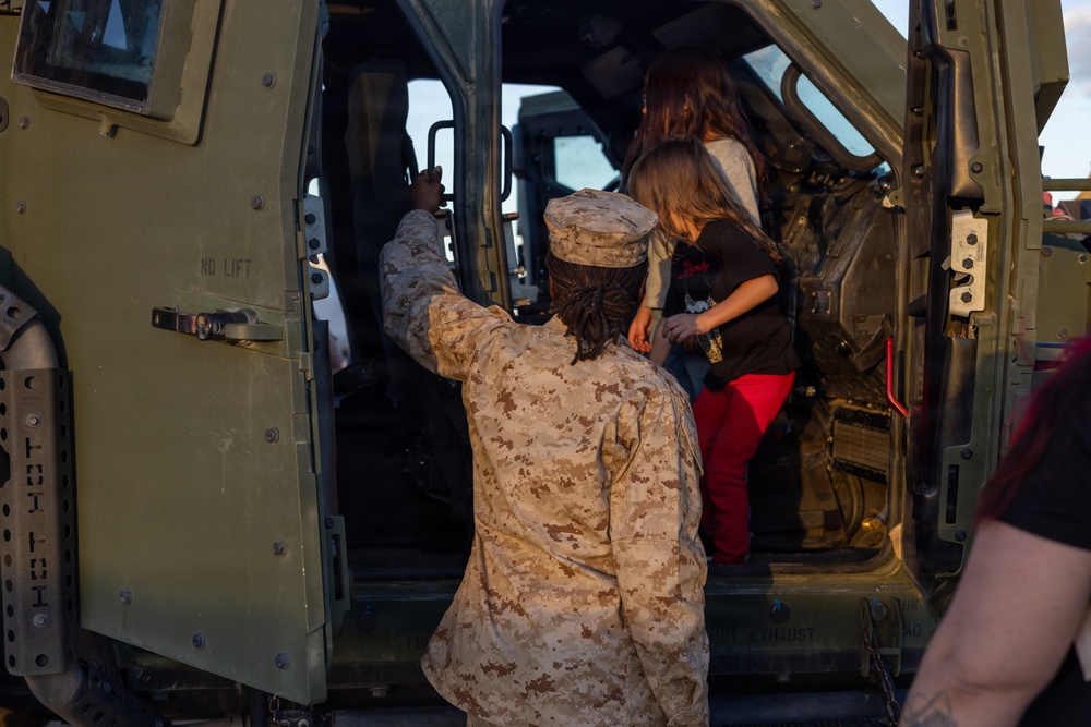 A U.S. Marine in desert MARPAT camouflage uniform assists a child and adult into a military vehicle, likely a HMMWV or similar tactical vehicle. The scene appears to be a public event or open house where civilians are being given access to military equipment. The Marine is providing hands-on assistance to help the child climb into the vehicle safely.