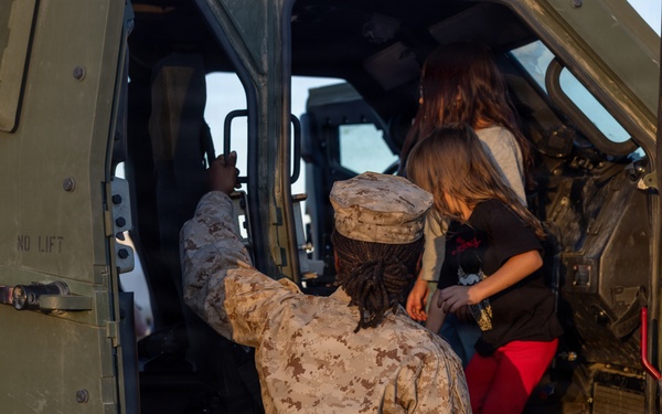 A U.S. Marine in desert MARPAT camouflage uniform assists a child and adult into a military vehicle, likely a HMMWV or similar tactical vehicle. The scene appears to be a public event or open house where civilians are being given access to military equipment. The Marine is providing hands-on assistance to help the child climb into the vehicle safely.