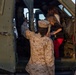 A U.S. Marine in desert MARPAT camouflage uniform assists a child and adult into a military vehicle, likely a HMMWV or similar tactical vehicle. The scene appears to be a public event or open house where civilians are being given access to military equipment. The Marine is providing hands-on assistance to help the child climb into the vehicle safely.