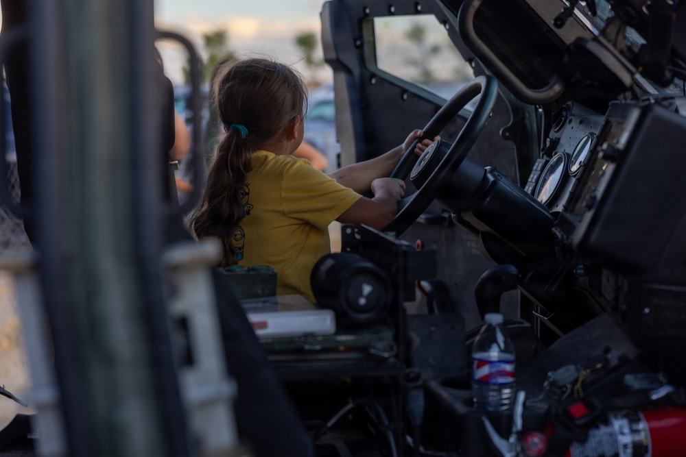 A young girl wearing a yellow t-shirt sits in the driver's seat of what appears to be a military vehicle, such as a Humvee, gripping the steering wheel. The setting appears to be an outdoor public event, likely a military open house or air show, given the dusk lighting and visible crowd in the background. The child appears engaged and excited to be exploring the vehicle.