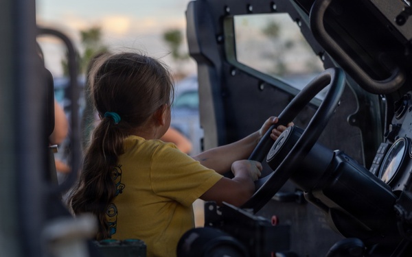 A young girl wearing a yellow t-shirt sits in the driver's seat of what appears to be a military vehicle, such as a Humvee, gripping the steering wheel. The setting appears to be an outdoor public event, likely a military open house or air show, given the dusk lighting and visible crowd in the background. The child appears engaged and excited to be exploring the vehicle.