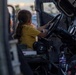 A young girl wearing a yellow t-shirt sits in the driver's seat of what appears to be a military vehicle, such as a Humvee, gripping the steering wheel. The setting appears to be an outdoor public event, likely a military open house or air show, given the dusk lighting and visible crowd in the background. The child appears engaged and excited to be exploring the vehicle.