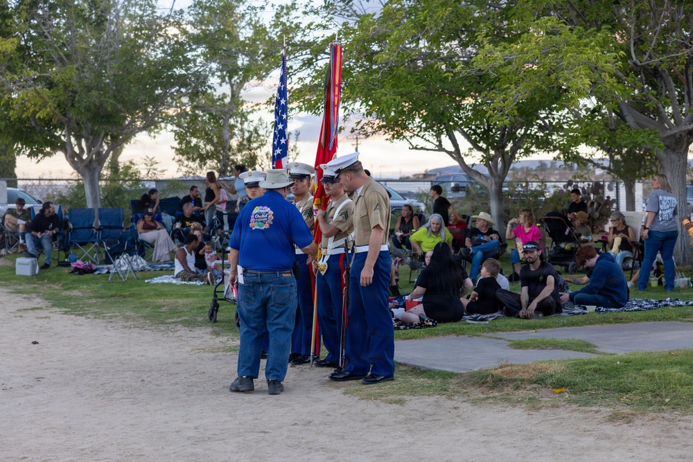 A color guard, appearing to be U.S. Marines in dress uniforms, is assembled at an outdoor public event in a park setting during evening hours. They are holding the American flag and what appears to be a Marine Corps flag, speaking with a civilian in a blue polo shirt. A large crowd of spectators is seated on the grass and in folding chairs in the background.