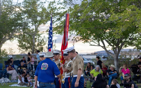 A color guard, appearing to be U.S. Marines in dress uniforms, is assembled at an outdoor public event in a park setting during evening hours. They are holding the American flag and what appears to be a Marine Corps flag, speaking with a civilian in a blue polo shirt. A large crowd of spectators is seated on the grass and in folding chairs in the background.