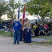 A color guard, appearing to be U.S. Marines in dress uniforms, is assembled at an outdoor public event in a park setting during evening hours. They are holding the American flag and what appears to be a Marine Corps flag, speaking with a civilian in a blue polo shirt. A large crowd of spectators is seated on the grass and in folding chairs in the background.