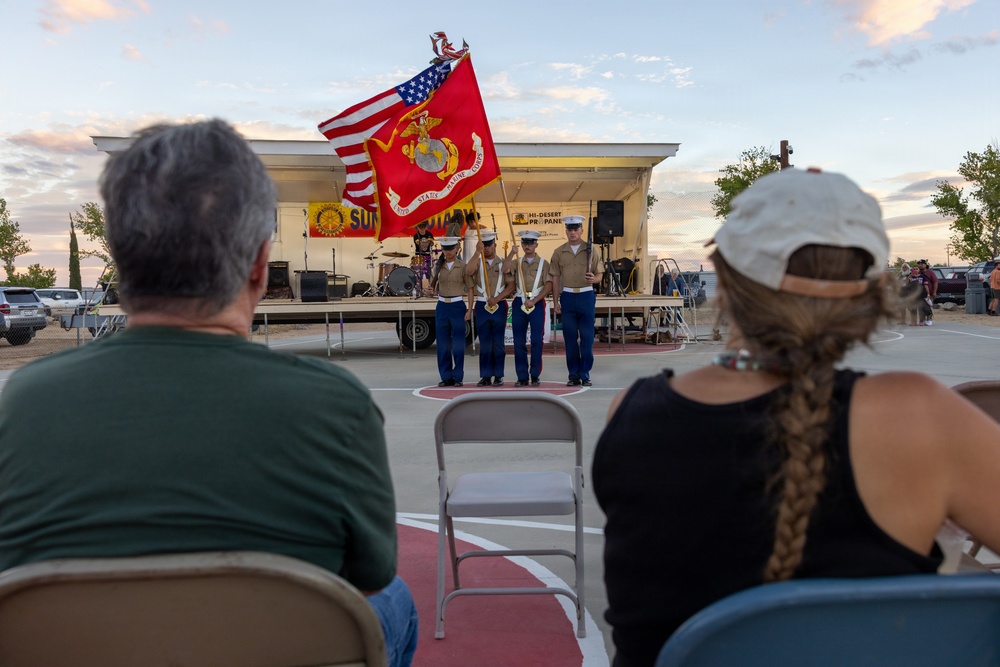 A United States Marine Corps color guard is presenting flags at an outdoor community event, likely a ceremony or public gathering at dusk. Four Marines in dress blues are holding the American flag and the Marine Corps flag in front of a small outdoor stage. Two civilian spectators are seated in the foreground watching the presentation.