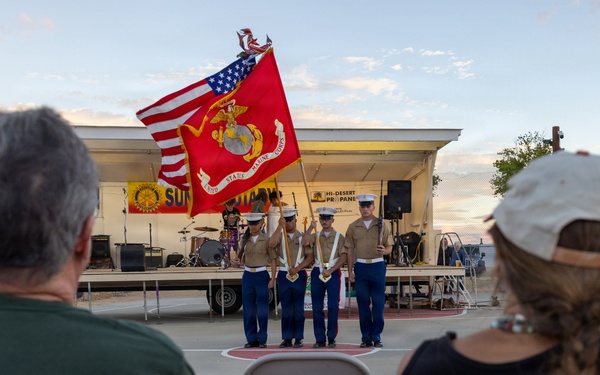 A United States Marine Corps color guard is presenting flags at an outdoor community event, likely a ceremony or public gathering at dusk. Four Marines in dress blues are holding the American flag and the Marine Corps flag in front of a small outdoor stage. Two civilian spectators are seated in the foreground watching the presentation.