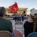 A United States Marine Corps color guard is presenting flags at an outdoor community event, likely a ceremony or public gathering at dusk. Four Marines in dress blues are holding the American flag and the Marine Corps flag in front of a small outdoor stage. Two civilian spectators are seated in the foreground watching the presentation.