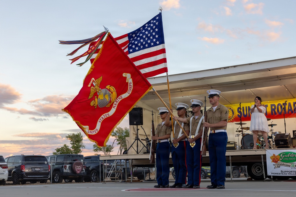 A United States Marine Corps color guard presents flags at an outdoor community event, likely a Rotary Club gathering based on visible signage. Four Marines in dress uniforms hold the USMC flag and the American flag, with two additional Marines carrying rifles. The ceremony takes place in front of a stage at dusk, with a woman in a white dress visible on the stage behind them.
