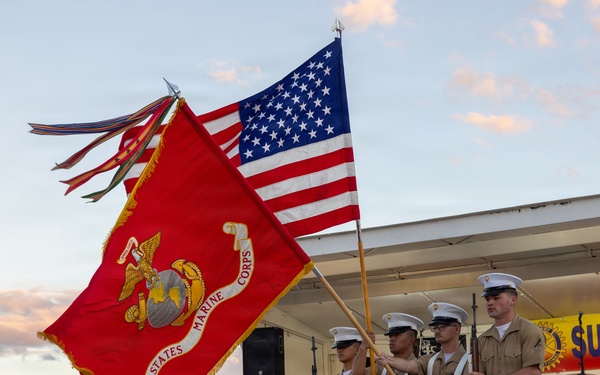A United States Marine Corps color guard presents flags at an outdoor community event, likely a Rotary Club gathering based on visible signage. Four Marines in dress uniforms hold the USMC flag and the American flag, with two additional Marines carrying rifles. The ceremony takes place in front of a stage at dusk, with a woman in a white dress visible on the stage behind them.