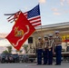 A United States Marine Corps color guard presents flags at an outdoor community event, likely a Rotary Club gathering based on visible signage. Four Marines in dress uniforms hold the USMC flag and the American flag, with two additional Marines carrying rifles. The ceremony takes place in front of a stage at dusk, with a woman in a white dress visible on the stage behind them.