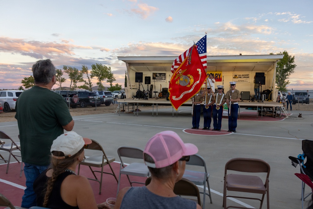 A United States Marine Corps color guard is presenting flags, including the American flag and the USMC flag, at an outdoor community event near a small stage. Audience members are seated in folding chairs watching the ceremony at dusk, with one man standing in apparent respect. The event appears to be taking place in a parking lot or outdoor venue with a performance stage visible in the background.