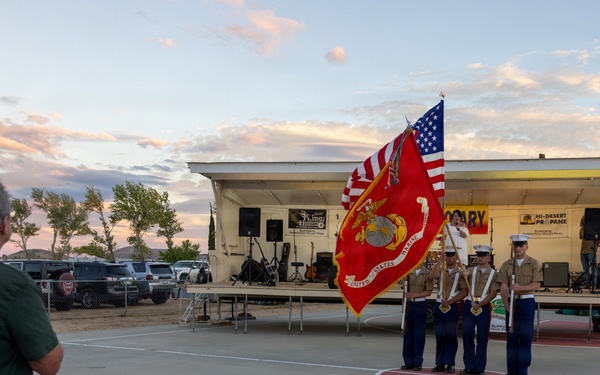 A United States Marine Corps color guard is presenting flags, including the American flag and the USMC flag, at an outdoor community event near a small stage. Audience members are seated in folding chairs watching the ceremony at dusk, with one man standing in apparent respect. The event appears to be taking place in a parking lot or outdoor venue with a performance stage visible in the background.