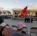 A United States Marine Corps color guard is presenting flags, including the American flag and the USMC flag, at an outdoor community event near a small stage. Audience members are seated in folding chairs watching the ceremony at dusk, with one man standing in apparent respect. The event appears to be taking place in a parking lot or outdoor venue with a performance stage visible in the background.