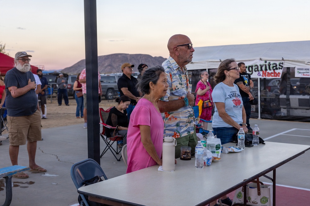 An outdoor community event or festival taking place at dusk in what appears to be a desert or arid region, with mountains visible in the background. Several people of varying ages are gathered around a folding table with water bottles and personal items. A food/drink vendor tent advertising 'Margaritas &amp; Nachos' is visible in the background.