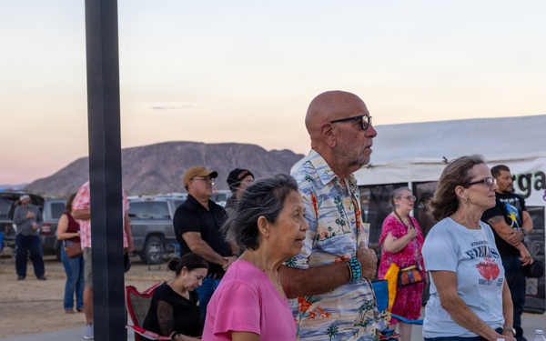 An outdoor community event or festival taking place at dusk in what appears to be a desert or arid region, with mountains visible in the background. Several people of varying ages are gathered around a folding table with water bottles and personal items. A food/drink vendor tent advertising 'Margaritas &amp; Nachos' is visible in the background.