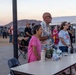 An outdoor community event or festival taking place at dusk in what appears to be a desert or arid region, with mountains visible in the background. Several people of varying ages are gathered around a folding table with water bottles and personal items. A food/drink vendor tent advertising 'Margaritas &amp; Nachos' is visible in the background.