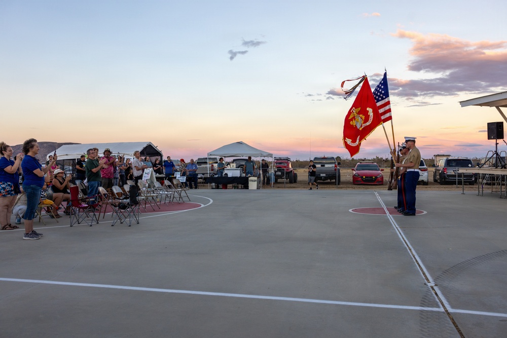 A U.S. Marine color guard ceremony is taking place on an outdoor concrete court at dusk, with two Marines holding the U.S. flag and the Marine Corps flag. A crowd of civilians stands and watches from the left side, many appearing to be at attention or taking photos. The setting appears to be a community or military-adjacent outdoor event in an arid, desert-like environment.