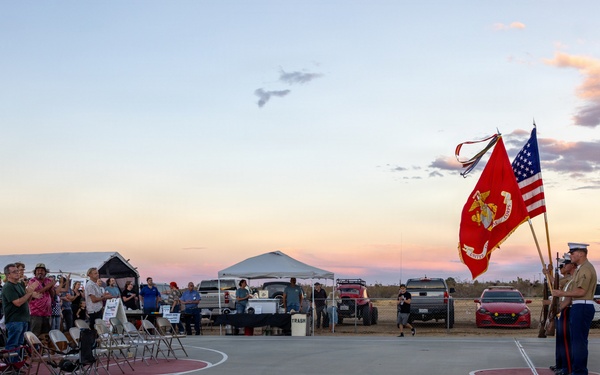 A U.S. Marine color guard ceremony is taking place on an outdoor concrete court at dusk, with two Marines holding the U.S. flag and the Marine Corps flag. A crowd of civilians stands and watches from the left side, many appearing to be at attention or taking photos. The setting appears to be a community or military-adjacent outdoor event in an arid, desert-like environment.
