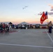 A U.S. Marine color guard ceremony is taking place on an outdoor concrete court at dusk, with two Marines holding the U.S. flag and the Marine Corps flag. A crowd of civilians stands and watches from the left side, many appearing to be at attention or taking photos. The setting appears to be a community or military-adjacent outdoor event in an arid, desert-like environment.