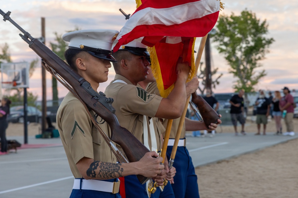 A U.S. Marine Corps color guard is performing a ceremonial presentation outdoors, likely at a community or military event. Marines in dress uniform are holding rifles and flags, including the American flag, in a formal formation. The event appears to be taking place at dusk on what looks like a recreational court or open plaza.