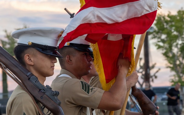 A U.S. Marine Corps color guard is performing a ceremonial presentation outdoors, likely at a community or military event. Marines in dress uniform are holding rifles and flags, including the American flag, in a formal formation. The event appears to be taking place at dusk on what looks like a recreational court or open plaza.