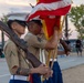 A U.S. Marine Corps color guard is performing a ceremonial presentation outdoors, likely at a community or military event. Marines in dress uniform are holding rifles and flags, including the American flag, in a formal formation. The event appears to be taking place at dusk on what looks like a recreational court or open plaza.