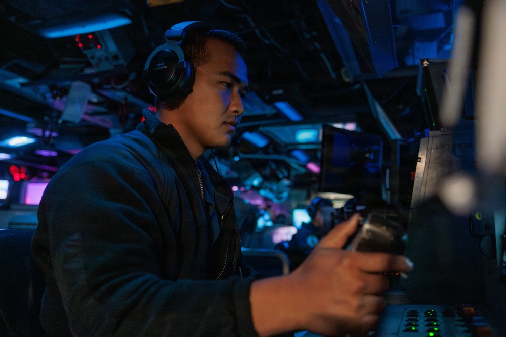A young male operator wearing over-ear headphones is seated at a control console in what appears to be a military naval vessel's combat information center or bridge. The environment is dimly lit with blue and red ambient lighting, characteristic of a ship's operational interior. He is actively manipulating a control device, likely a joystick or throttle, while monitoring equipment.