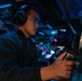 A young male operator wearing over-ear headphones is seated at a control console in what appears to be a military naval vessel's combat information center or bridge. The environment is dimly lit with blue and red ambient lighting, characteristic of a ship's operational interior. He is actively manipulating a control device, likely a joystick or throttle, while monitoring equipment.