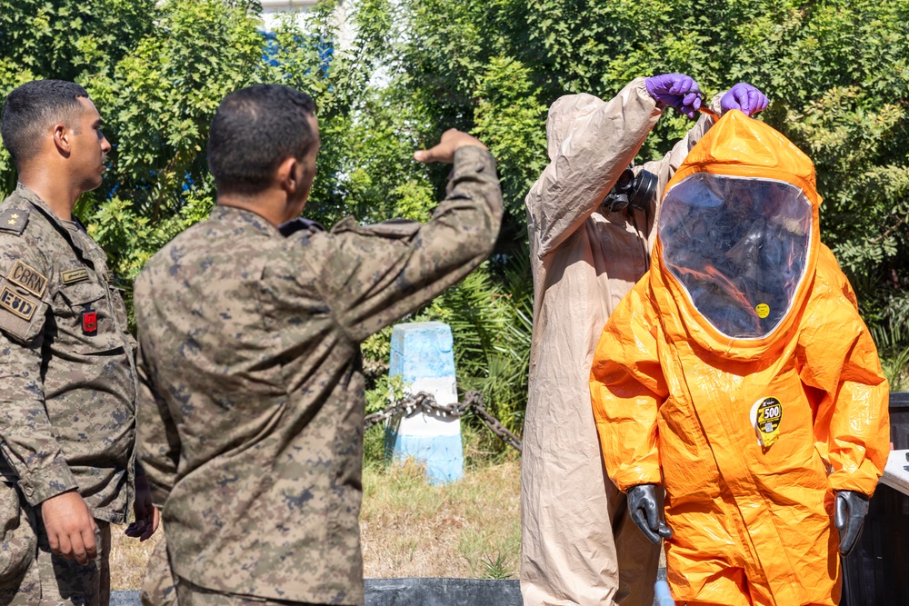 Military personnel in camouflage uniforms are assisting a colleague in donning or inspecting an orange Level A hazmat encapsulation suit with an integrated full-face respirator. The patch on the left soldier's uniform reads 'CBRN' and 'EUD', indicating Chemical, Biological, Radiological, and Nuclear specialization. The scene appears to be an outdoor training or demonstration exercise.