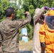 Military personnel in camouflage uniforms are assisting a colleague in donning or inspecting an orange Level A hazmat encapsulation suit with an integrated full-face respirator. The patch on the left soldier's uniform reads 'CBRN' and 'EUD', indicating Chemical, Biological, Radiological, and Nuclear specialization. The scene appears to be an outdoor training or demonstration exercise.