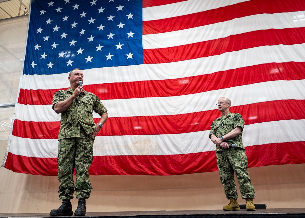 Two U.S. Navy personnel in NWU Type III (green camouflage) uniforms stand on a raised stage in front of a large American flag. The individual on the left is speaking into a microphone while the one on the right stands at ease. The setting appears to be an indoor military facility, likely a hangar or large assembly hall.
