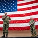 Two U.S. Navy personnel in NWU Type III (green camouflage) uniforms stand on a raised stage in front of a large American flag. The individual on the left is speaking into a microphone while the one on the right stands at ease. The setting appears to be an indoor military facility, likely a hangar or large assembly hall.