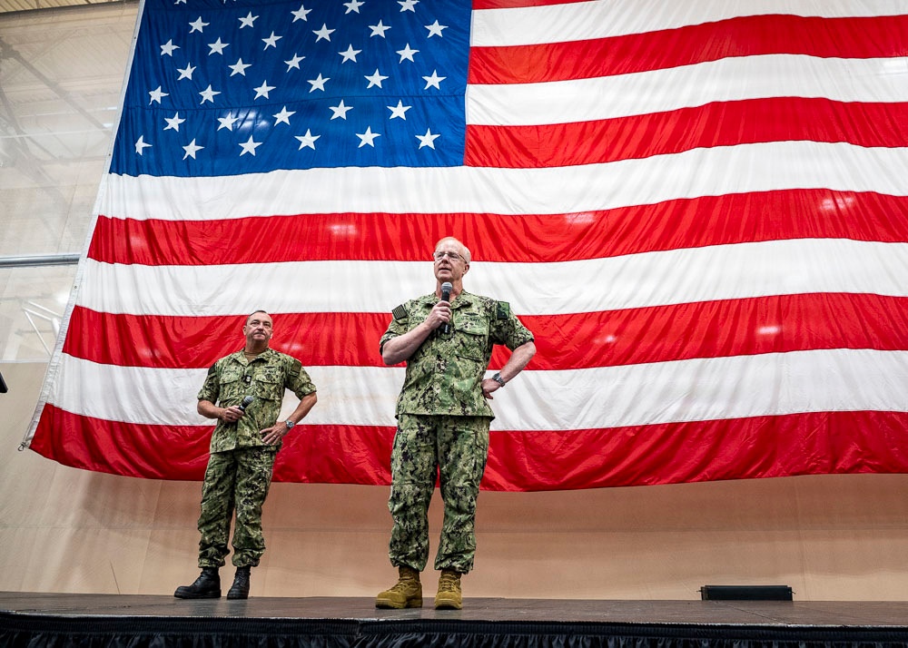 Two U.S. Navy personnel in camouflage uniforms stand on a stage in front of a large American flag. The taller individual on the right is speaking into a microphone while the other stands at ease to the left. The setting appears to be an indoor military event or all-hands briefing.
