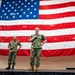 Two U.S. Navy personnel in camouflage uniforms stand on a stage in front of a large American flag. The taller individual on the right is speaking into a microphone while the other stands at ease to the left. The setting appears to be an indoor military event or all-hands briefing.
