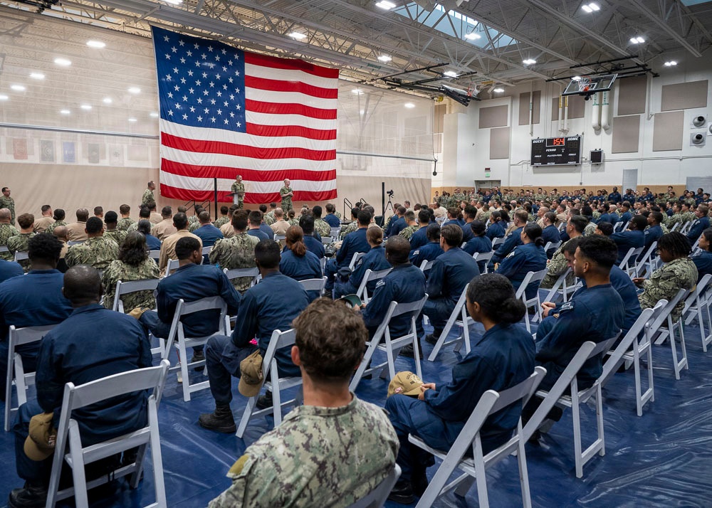 A large military assembly is taking place inside a gymnasium or indoor sports facility, with hundreds of uniformed personnel seated in white folding chairs facing a speaker at a podium. A large American flag is prominently displayed on the wall behind the speaker, serving as a backdrop for what appears to be an official military briefing or ceremony. The attendees are predominantly wearing Navy Working Uniform (NWU) blue digital camouflage.