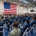 A large military assembly is taking place inside a gymnasium or indoor sports facility, with hundreds of uniformed personnel seated in white folding chairs facing a speaker at a podium. A large American flag is prominently displayed on the wall behind the speaker, serving as a backdrop for what appears to be an official military briefing or ceremony. The attendees are predominantly wearing Navy Working Uniform (NWU) blue digital camouflage.
