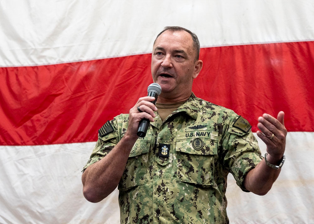 A U.S. Navy officer in digital camouflage uniform (NWU Type III) is speaking into a handheld microphone, appearing to address an audience. He is standing in front of a large American flag used as a backdrop. His uniform displays 'U.S. NAVY' tape, various insignia, and rank indicators consistent with a senior enlisted or officer grade.