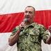 A U.S. Navy officer in digital camouflage uniform (NWU Type III) is speaking into a handheld microphone, appearing to address an audience. He is standing in front of a large American flag used as a backdrop. His uniform displays 'U.S. NAVY' tape, various insignia, and rank indicators consistent with a senior enlisted or officer grade.