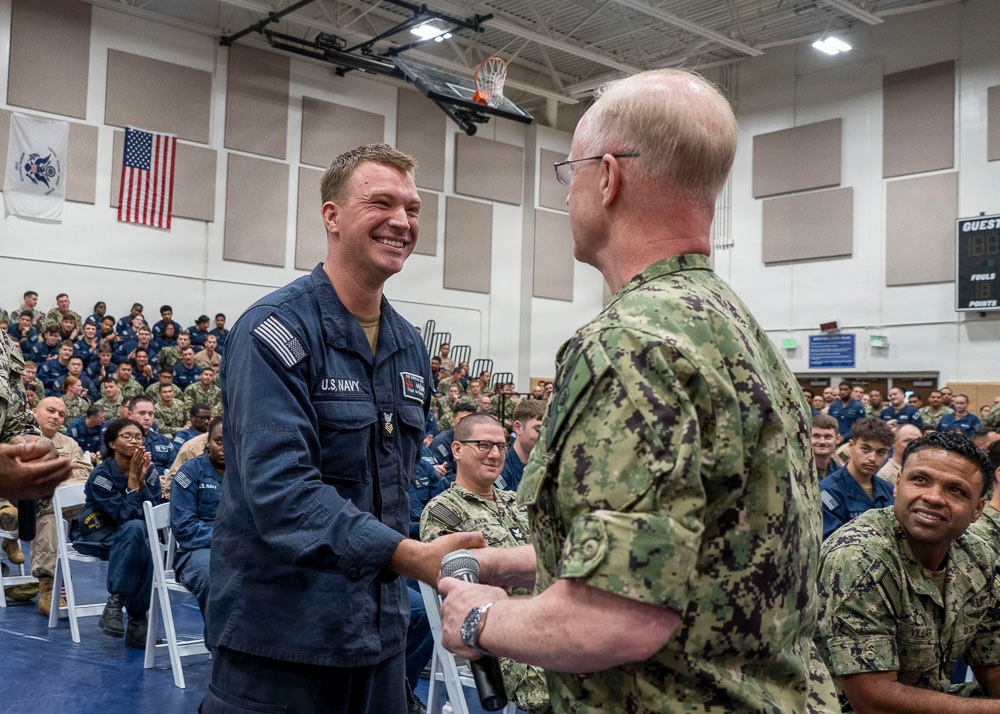 A U.S. Navy ceremony taking place inside a gymnasium or multipurpose facility, where a senior officer in camouflage NWU (Navy Working Uniform) Type III is shaking hands with an enlisted sailor in Navy blue coveralls. The sailor is smiling broadly, suggesting a recognition or award event. A large audience of military personnel in various uniforms is seated in the background observing the event.