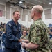 A U.S. Navy ceremony taking place inside a gymnasium or multipurpose facility, where a senior officer in camouflage NWU (Navy Working Uniform) Type III is shaking hands with an enlisted sailor in Navy blue coveralls. The sailor is smiling broadly, suggesting a recognition or award event. A large audience of military personnel in various uniforms is seated in the background observing the event.