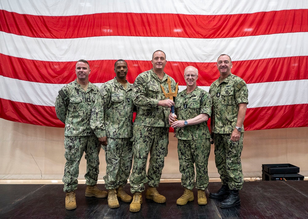 Five U.S. Navy personnel in NWU Type III (AOR-2) camouflage uniforms are posed for a formal group photo in front of a large American flag backdrop. The two center individuals are jointly holding what appears to be a wooden trident-shaped award or plaque, suggesting a recognition or award ceremony. The setting appears to be an indoor military facility or hangar.