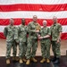 Five U.S. Navy personnel in NWU Type III (AOR-2) camouflage uniforms are posed for a formal group photo in front of a large American flag backdrop. The two center individuals are jointly holding what appears to be a wooden trident-shaped award or plaque, suggesting a recognition or award ceremony. The setting appears to be an indoor military facility or hangar.
